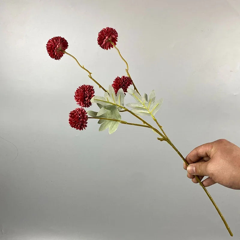 Una ramita de flores artificiales de crisantemo rojo con hojas verdes sostenida por una mano contra un fondo gris claro.
