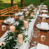 A long wooden table set for an outdoor event with white linens, glassware, and pink candles in glass holders.