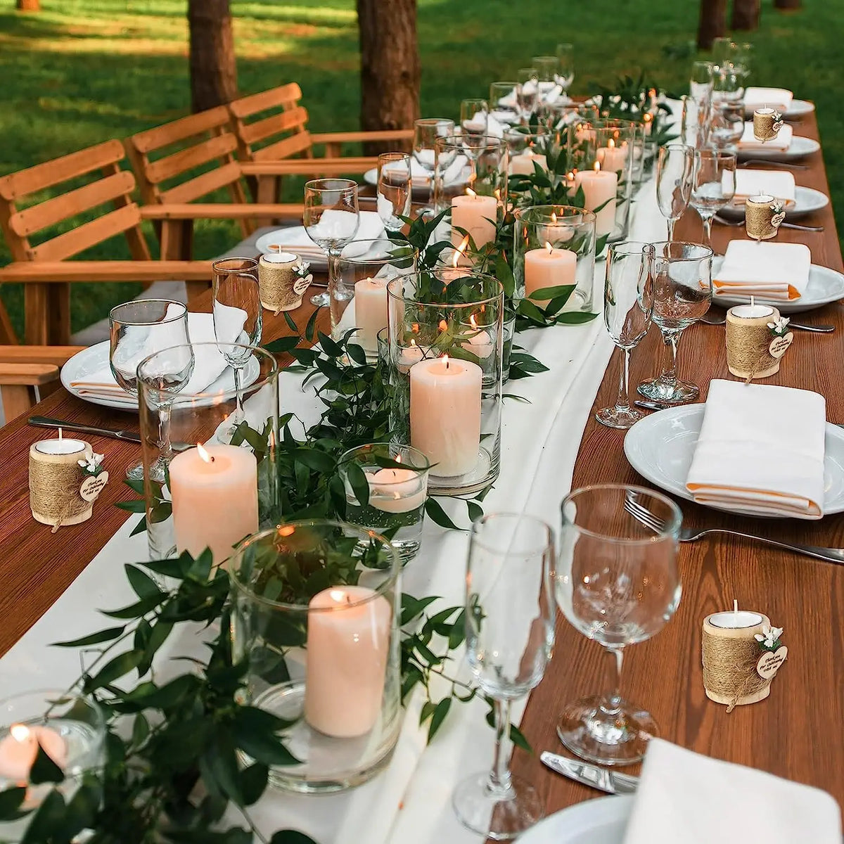 A long wooden table set for an outdoor event with white linens, glassware, and pink candles in glass holders.