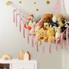 A multi-use storage hammock made of pink and white fabric hangs on a wall, filled with various stuffed animals and plush toys