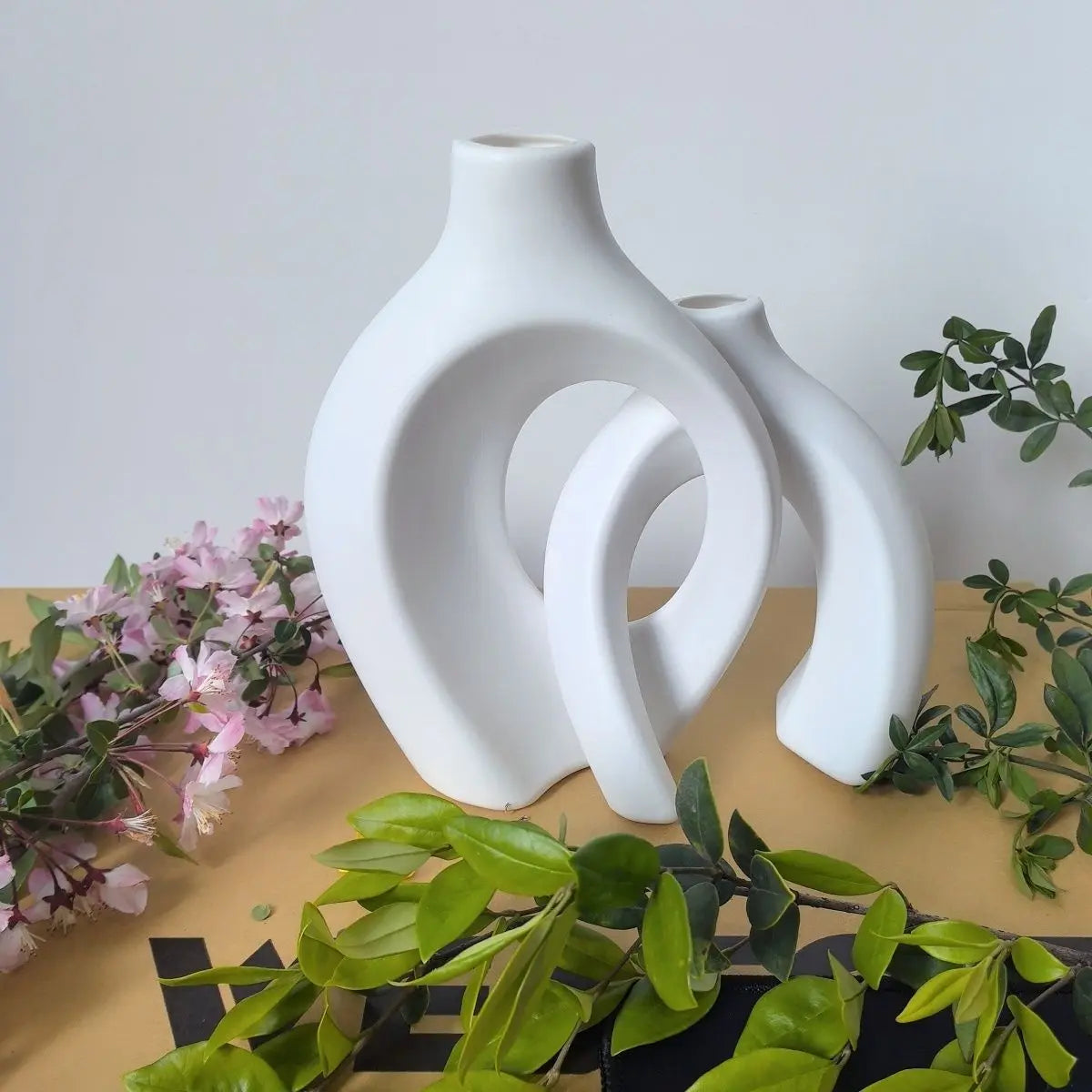 A set of two white, organic-shaped ceramic vases for home decor displayed on a wooden surface with greenery.