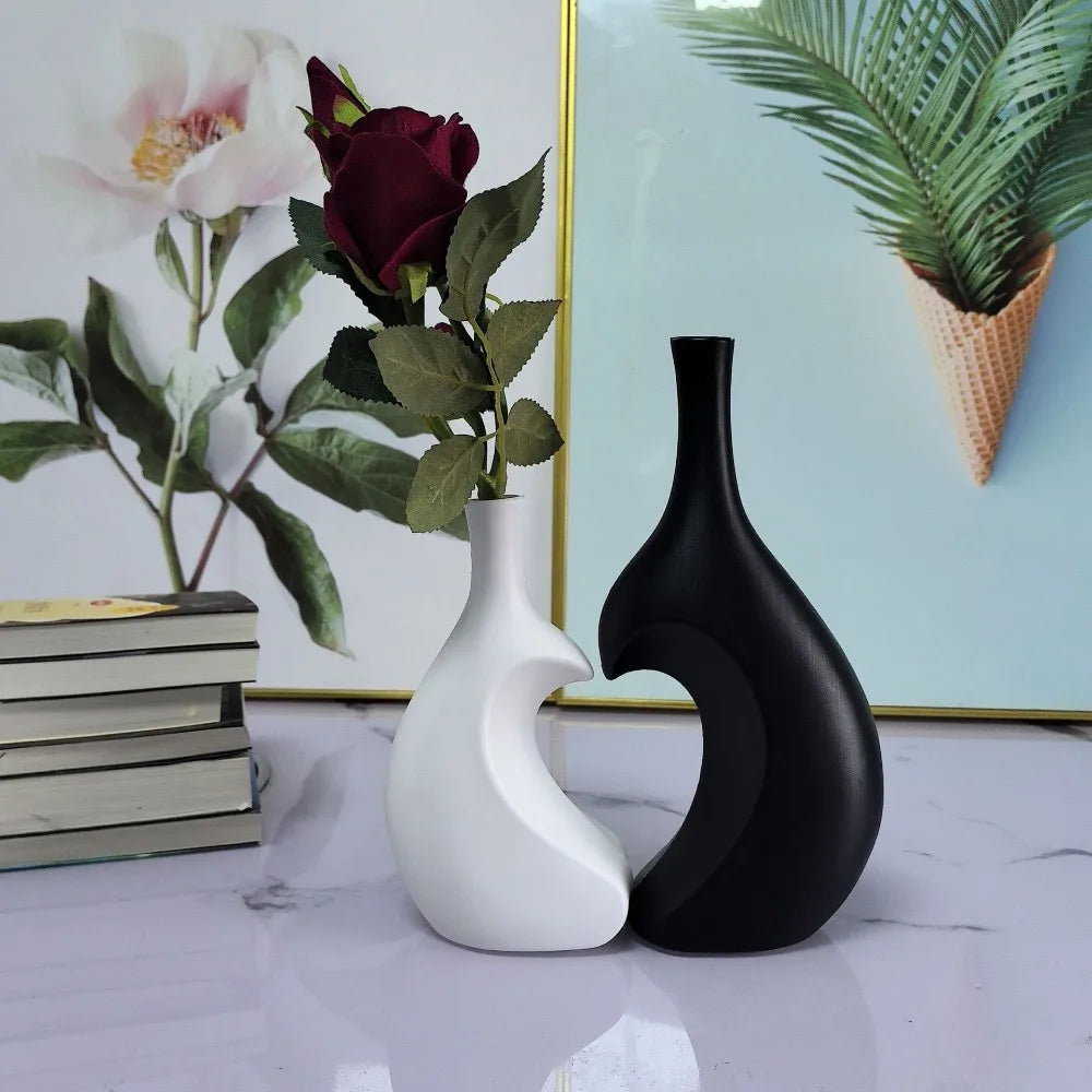 A modern heart-shaped ceramic vase in matte black and white sits on a marble surface next to a floral arrangement.