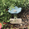 A light blue flower with a small bee on a wooden tag is placed in dark soil amidst green foliage.