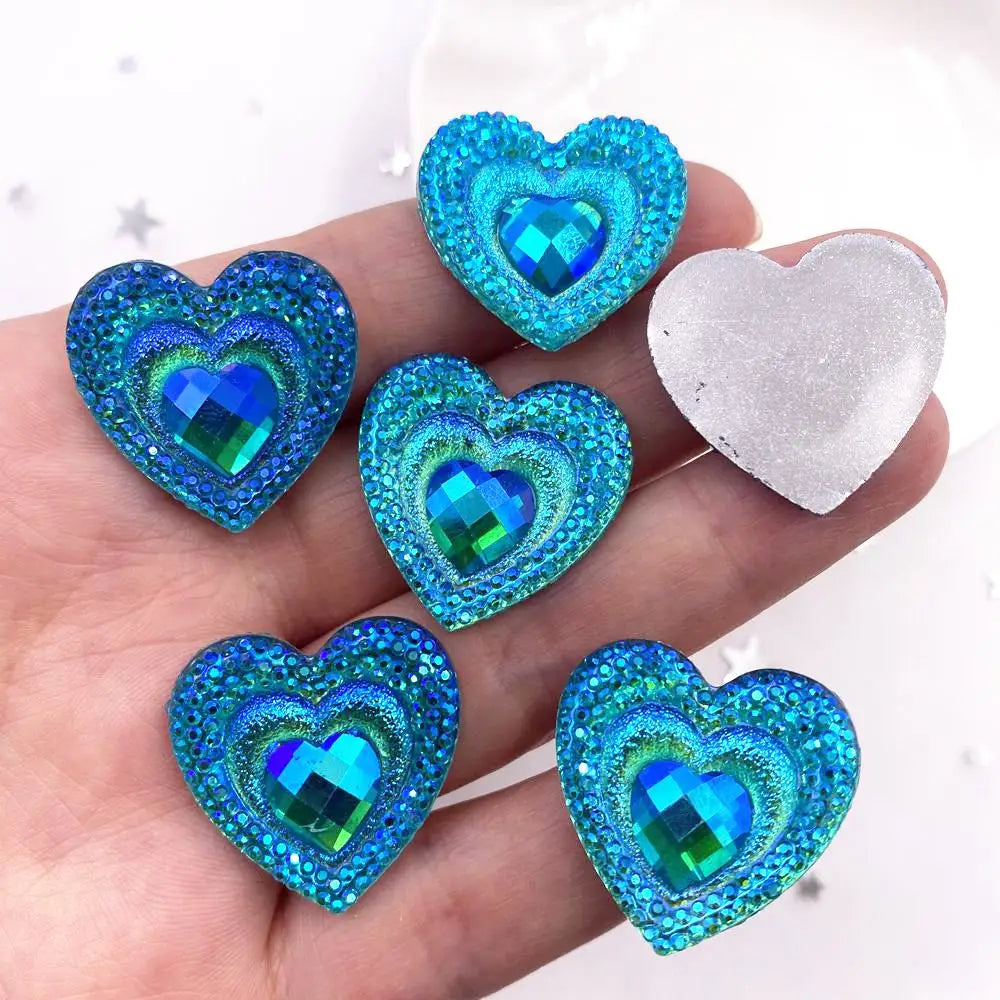 A hand holds a set of twelve heart-shaped blue iridescent peacock resin rhinestone flatbacks.