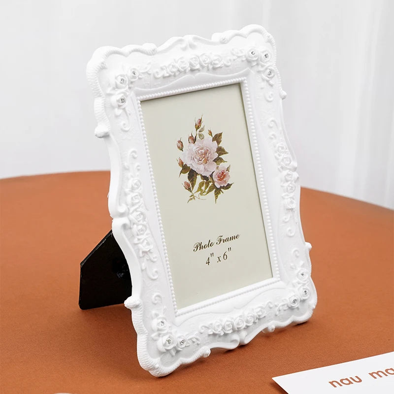 A white ornate frame with a distressed edge holds a pale pink rose floral design on a light background.