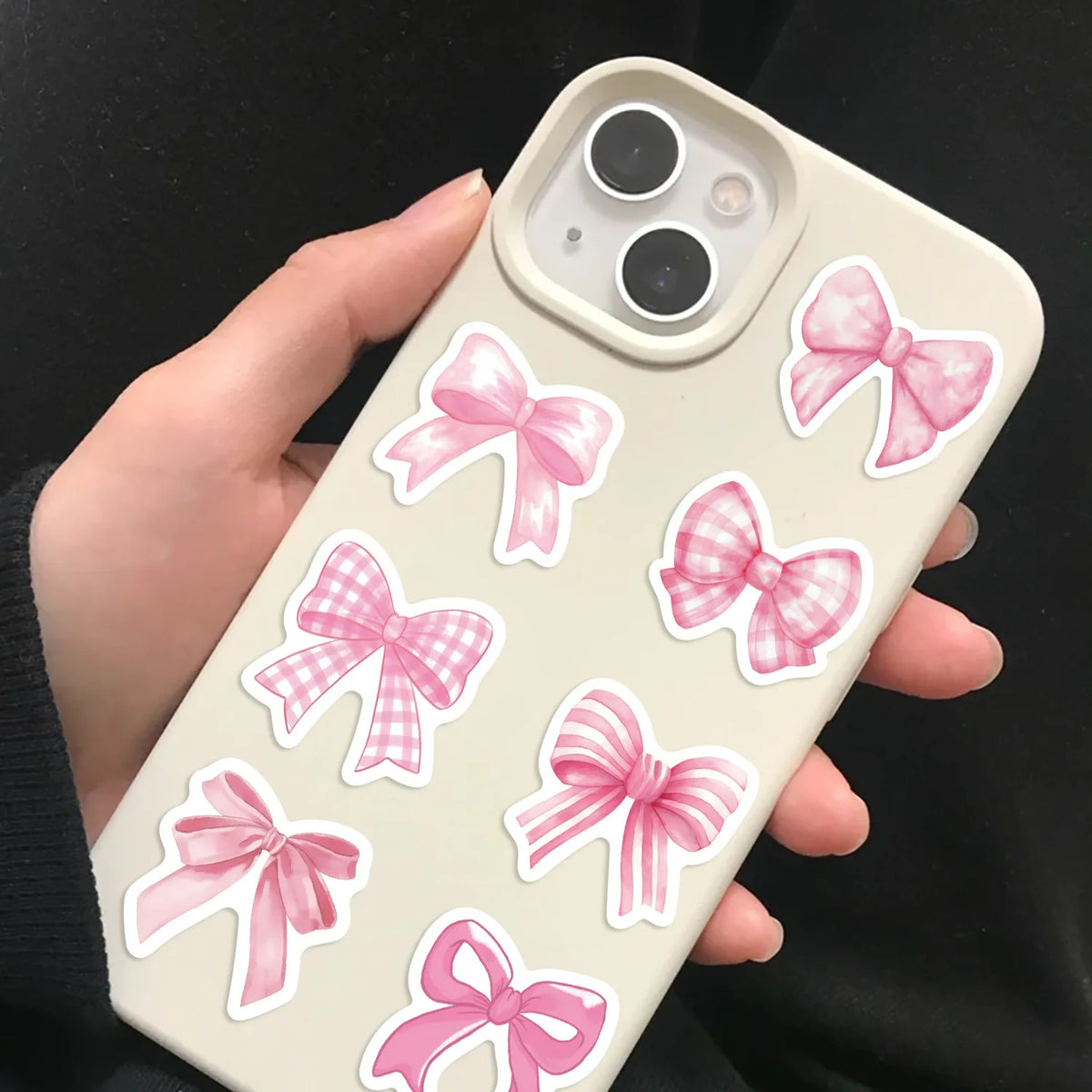 A hand holds a phone case decorated with pink bow stickers on a beige background.