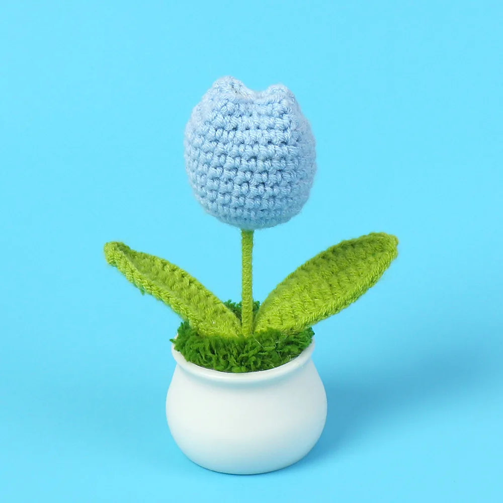 A hand-woven crochet blue flower with green leaves planted in a white pot.
