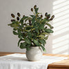 An artificial olive tree branch with green leaves and dark berries in a light gray ceramic pot.
