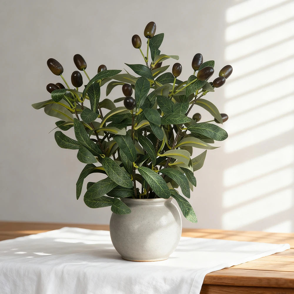 An artificial olive tree branch with green leaves and dark berries in a light gray ceramic pot.