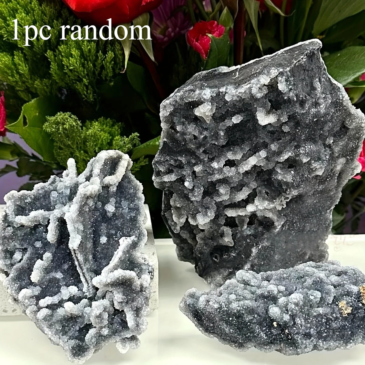 A cluster of dark gray natural zinc crystals displayed against a blurred background of green and red foliage.