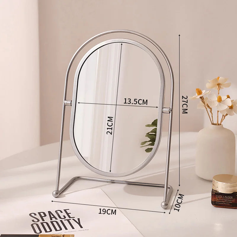A silver-framed oval vanity mirror with a rotating stand is displayed on a white surface.