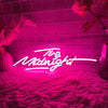 A neon sign spelling out The Midnight glowing bright pink against a textured pink rug and tiled wall.