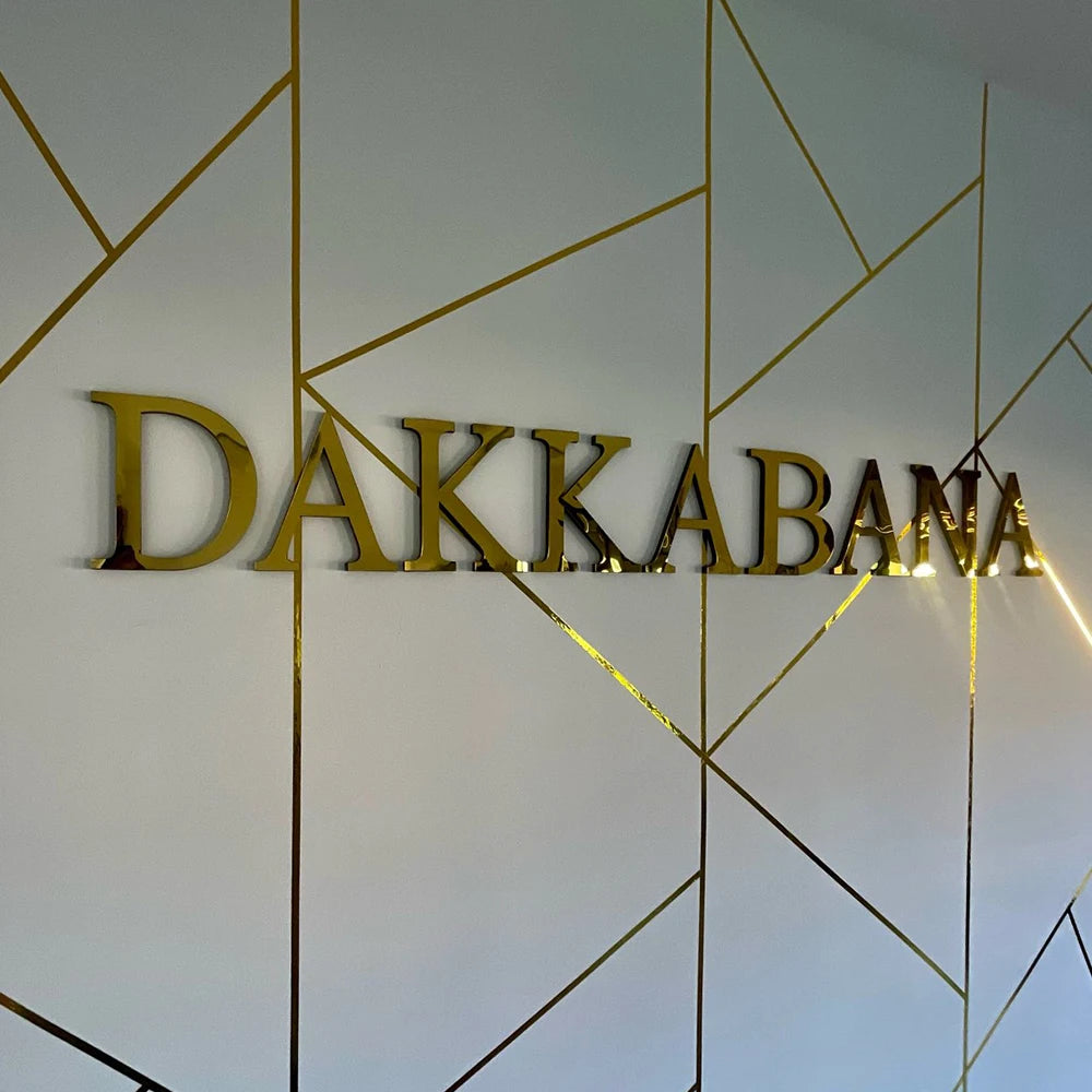 Gold acrylic mirror letter wall stickers spelling DAKKABANA on a white wall with geometric lines.