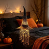 A glowing witch hat hanging chime with star tassels illuminates a dark bedroom setting.