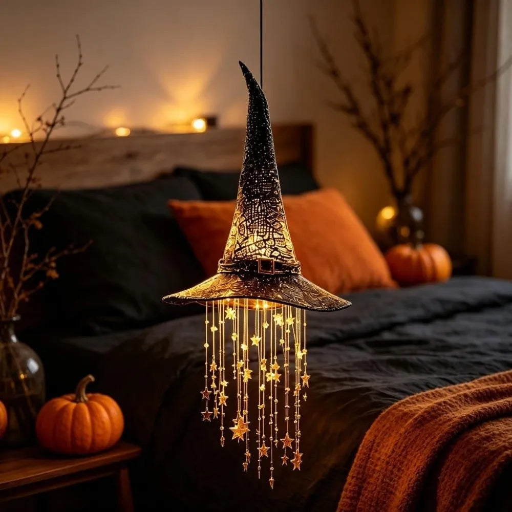 A glowing witch hat hanging chime with star tassels illuminates a dark bedroom setting.