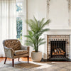 An artificial triple trunk palm tree in a beige pot stands in a bright living room next to a fireplace.