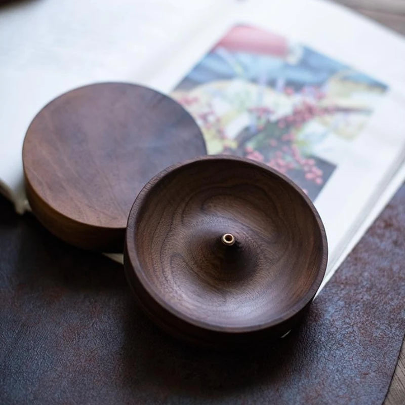 A dark brown solid wood incense stick burner with a round lid resting on a book.