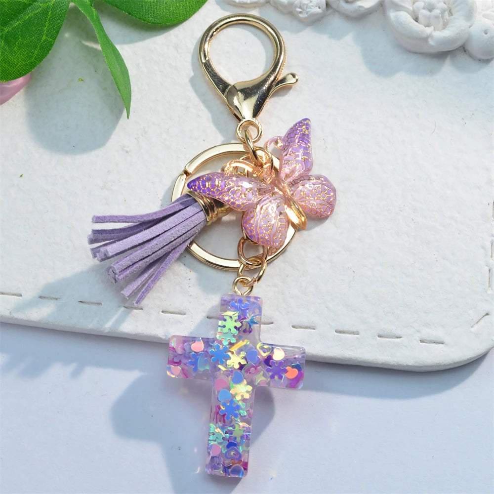 A gold-toned keychain featuring a purple tassel and a pink glitter butterfly charm with a purple resin cross.