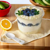 A glass of creamy blueberry pudding served in a clear plastic cup on a wooden board with an orange slice and green peppers ne