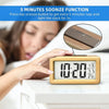 A hand presses a snooze button on a wooden digital alarm clock displaying 10:20, with a woman sleeping in the background.