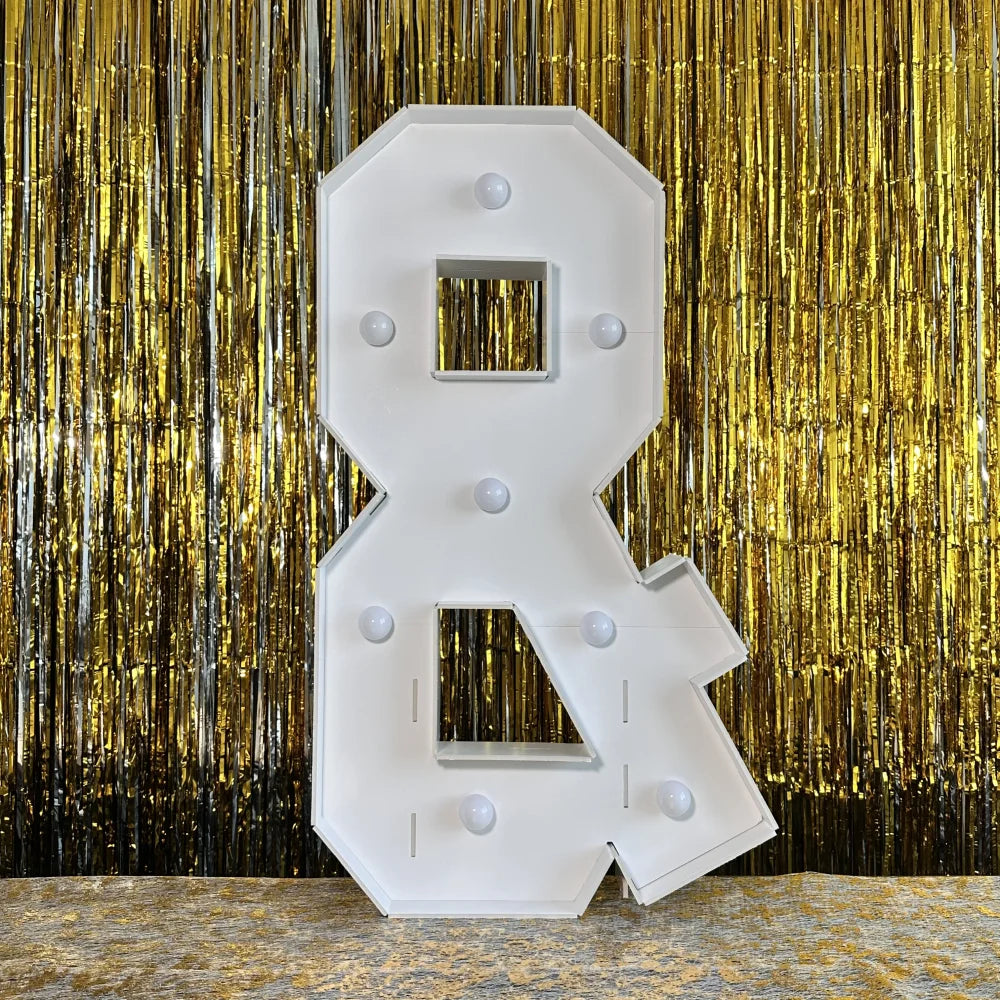 A large white marquee light-up letter for party decor is displayed against a shimmering gold curtain backdrop.
