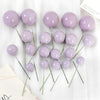 A set of twenty light purple spherical cake toppers on thin green stems arranged on a white cloth.