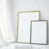 A framed piece of white art or print with a gold frame and a separate black frame leaning against a white wall.