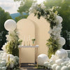 A cream-colored arch backdrop decorated with white floral greenery and round white balloons.