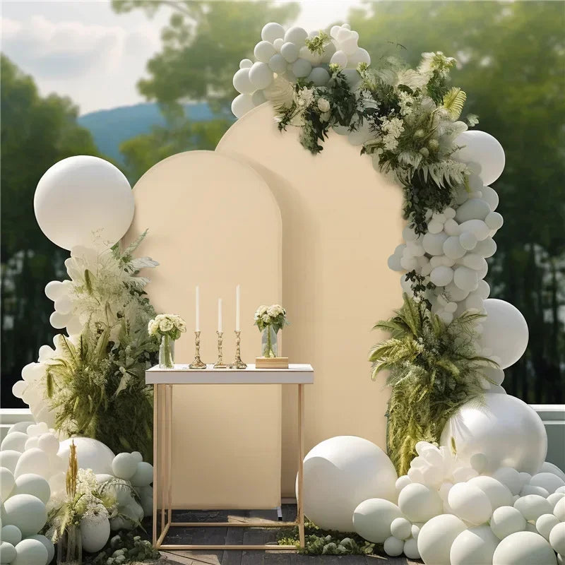 A cream-colored arch backdrop decorated with white floral greenery and round white balloons.