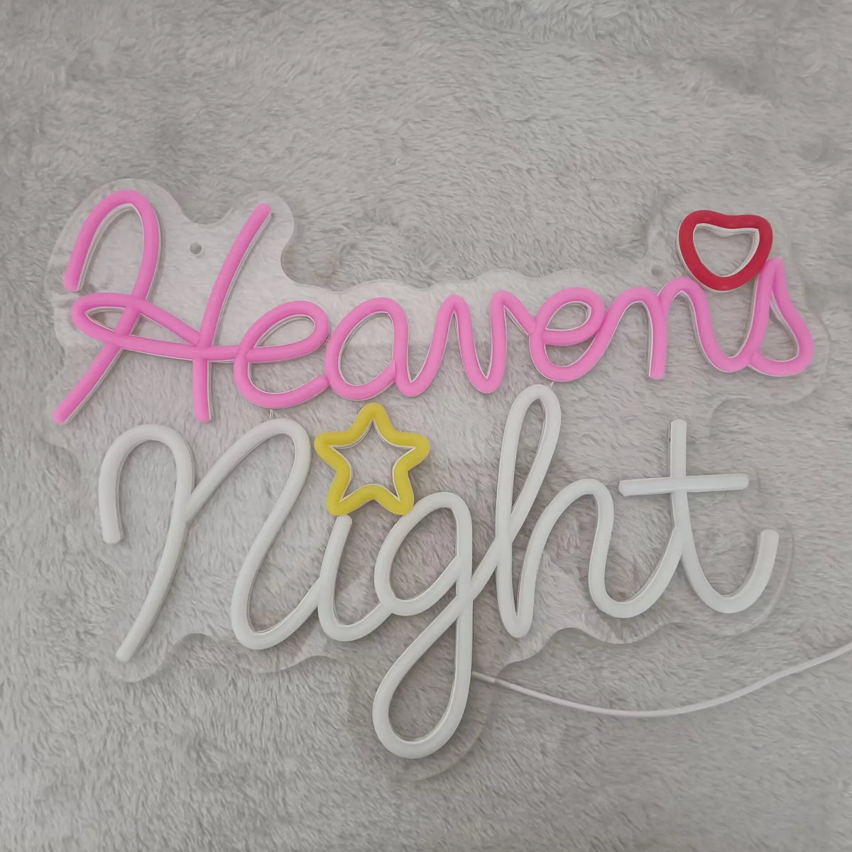 Pink and white neon sign spelling out Heavens Night on a fuzzy gray surface.