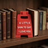 A red 3D printed letter sign ornament shaped like a house with the text I love you carved into it, displayed on a wooden shel