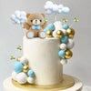 A white cake decorated with blue and gold round balls, featuring a brown bear topper and star decorations.