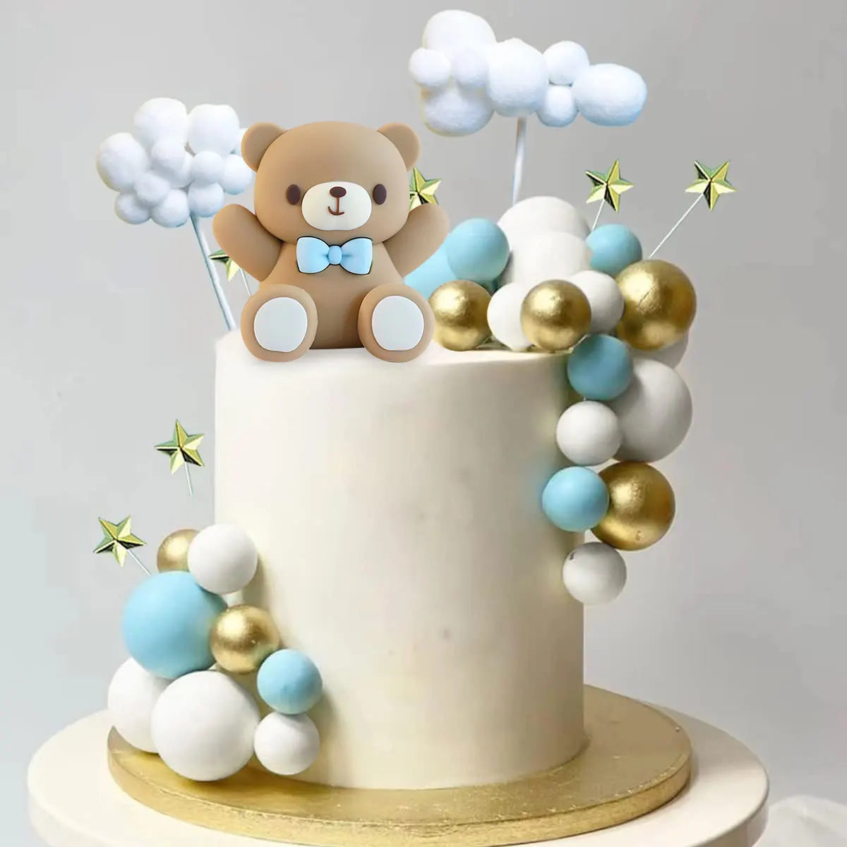 A white cake decorated with blue and gold round balls, featuring a brown bear topper and star decorations.