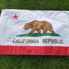 A white flag featuring a brown bear silhouette and a red star, with the text CALIFORNIA REPUBLIC printed below, laid out on g