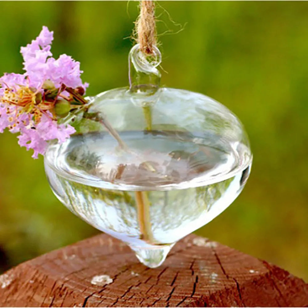 A clear glass hanging planter vase filled with water, suspended by twine, set against a blurred green background.