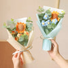 A hand holds a bouquet of handcrafted crochet sunflowers and greenery wrapped in light blue paper.