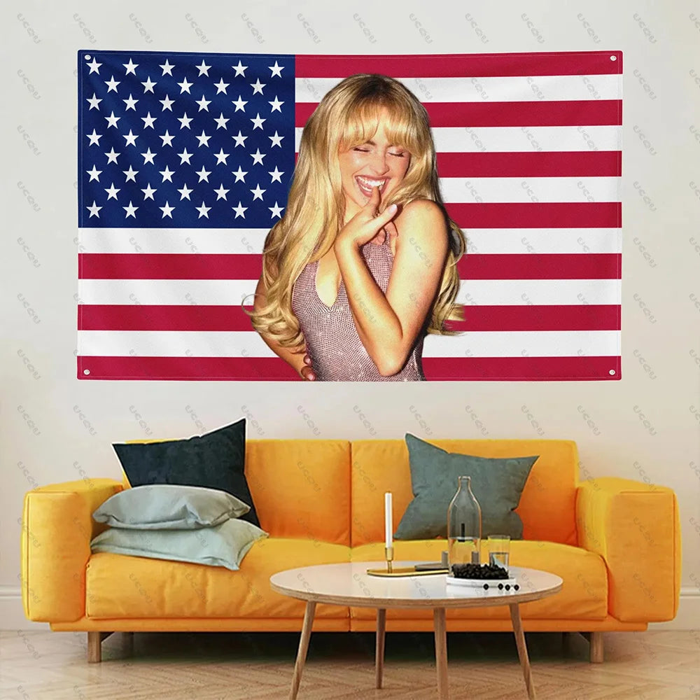 A woman with blonde hair is posing in front of an American flag tapestry decorated with red and white stripes and blue stars.