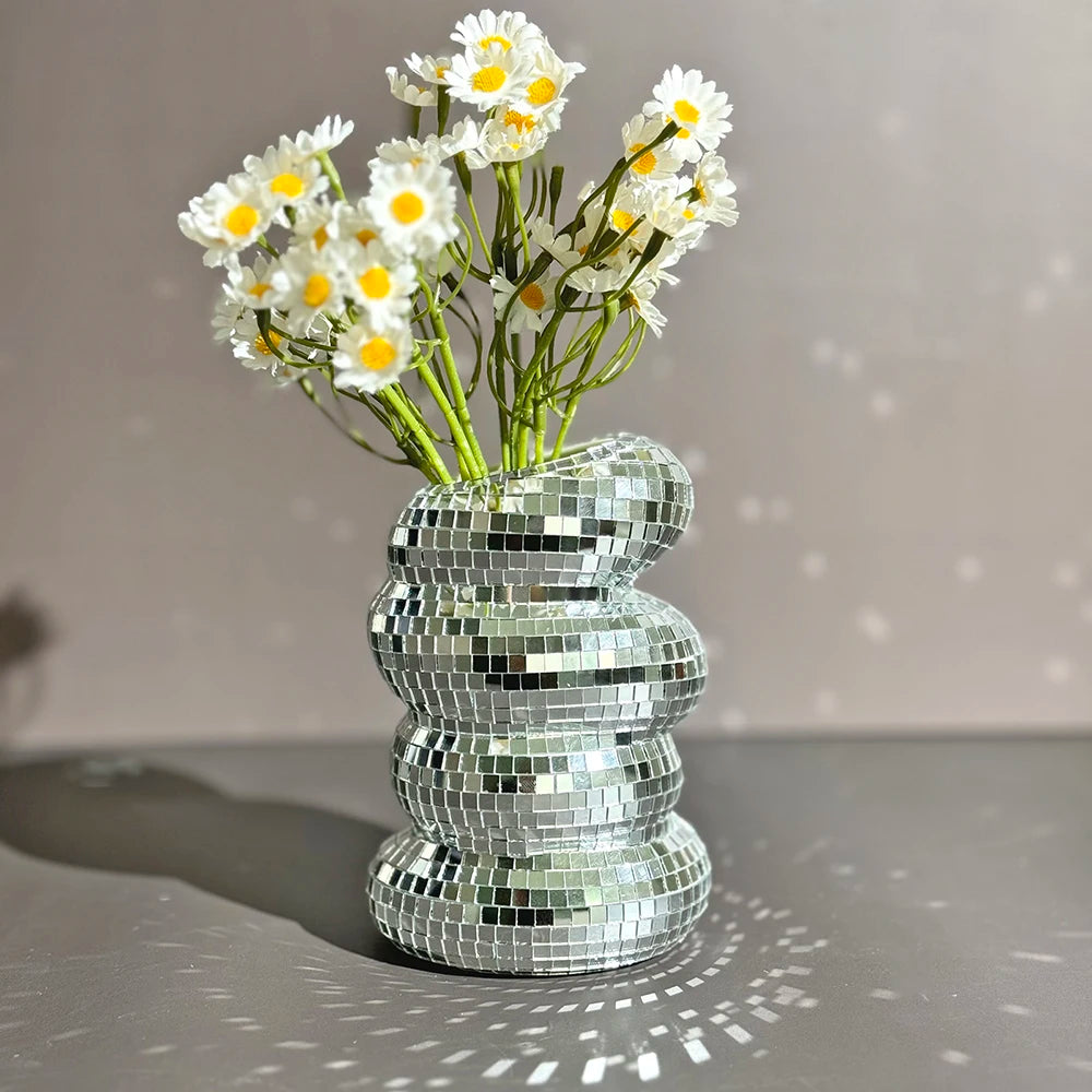 A vase filled with white daisies sits on a surface, surrounded by stacked silver mirror tiles.
