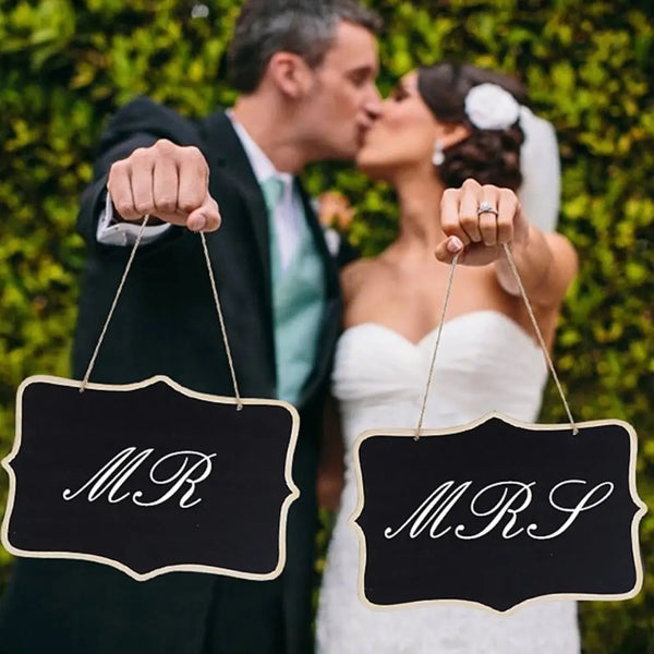 A bride and groom kiss while holding rustic wooden message boards for a wedding event.