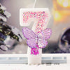 A white birthday candle shaped like the number 7 decorated with pink and purple glitter, set on a white surface.