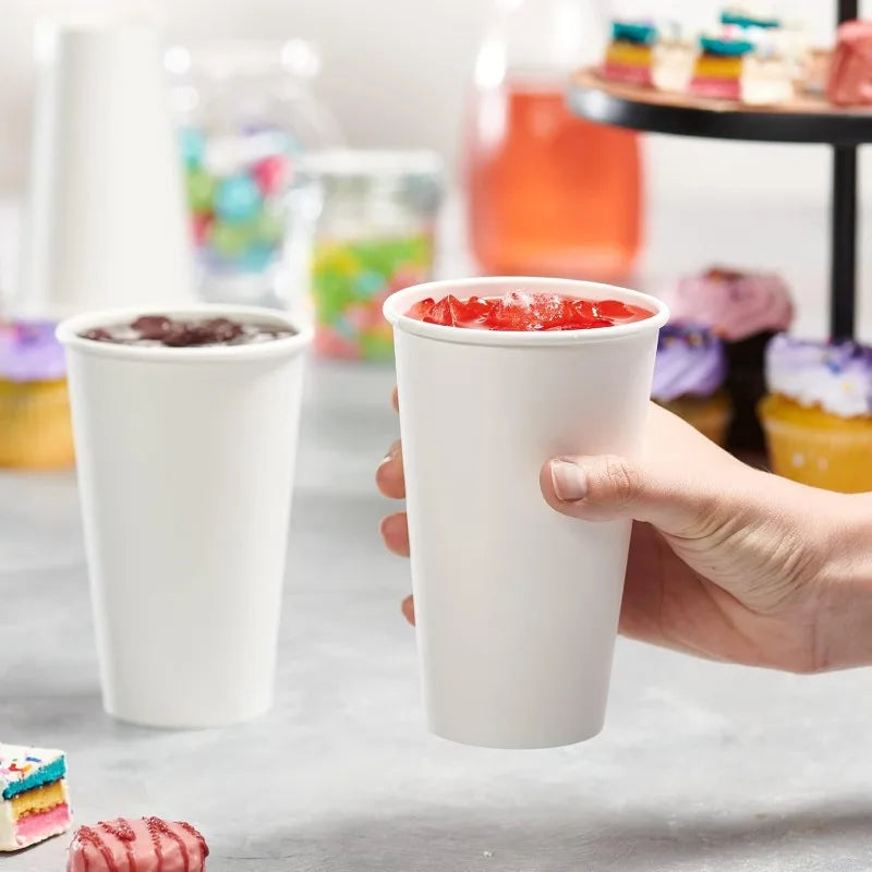 A hand holds a white disposable paper coffee cup filled with red fruit, set against a blurred background of colorful drinks.