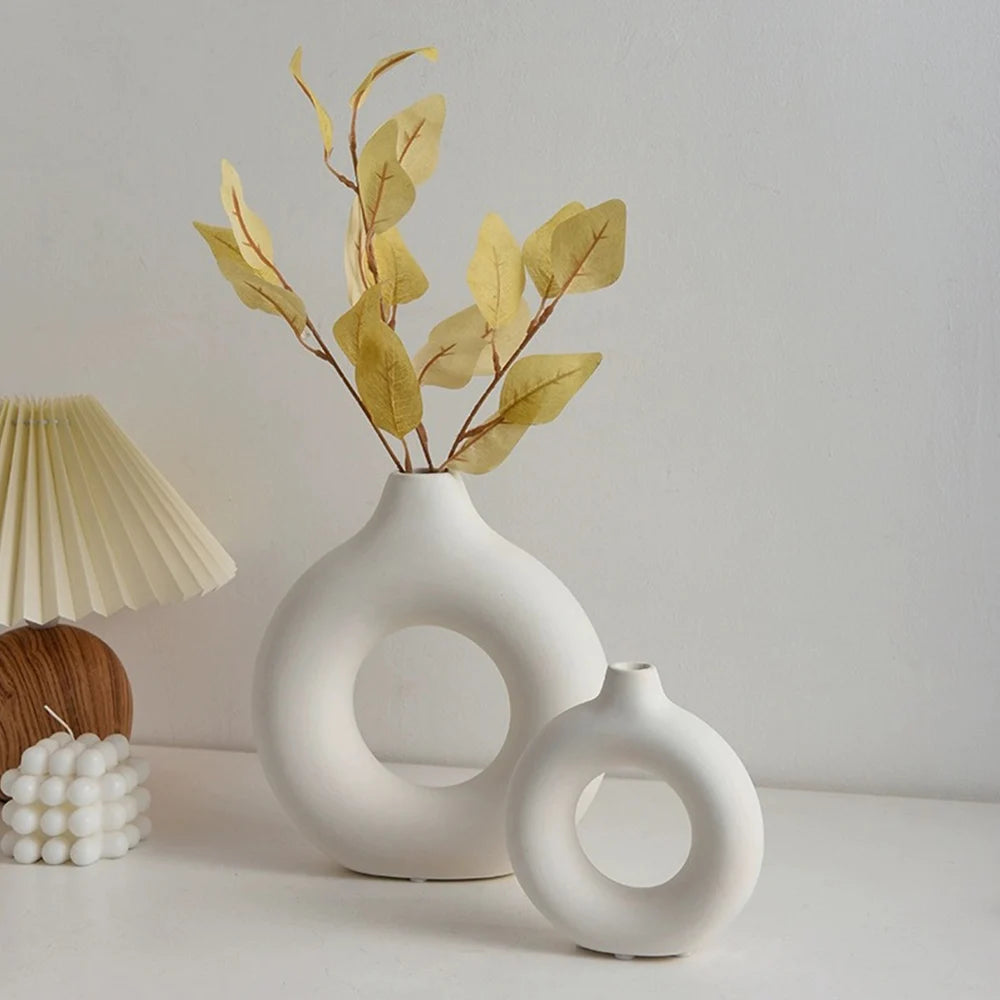 A white ceramic circular vase holds dried yellow leaves against a light background.