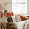 A large matte black decorative vase filled with orange and red flowers sits on a wooden table next to a bright bedroom window