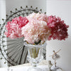 A close-up of pink and purple silk hydrangea stems with vintage burnt edge petals arranged in glass vases.