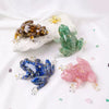 A collection of resin crystal frogs in various colors including amber, green, blue, and pink resting on a white rock.