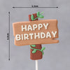 A wooden sign with 'HAPPY BIRTHDAY' written on it, featuring green leaves and fruit accents.