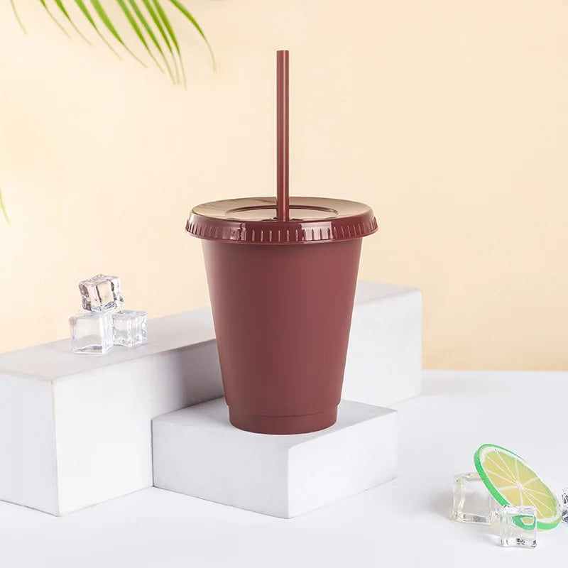 A brown plastic cold cup with a lid and straw is displayed on a white surface next to some ice cubes.