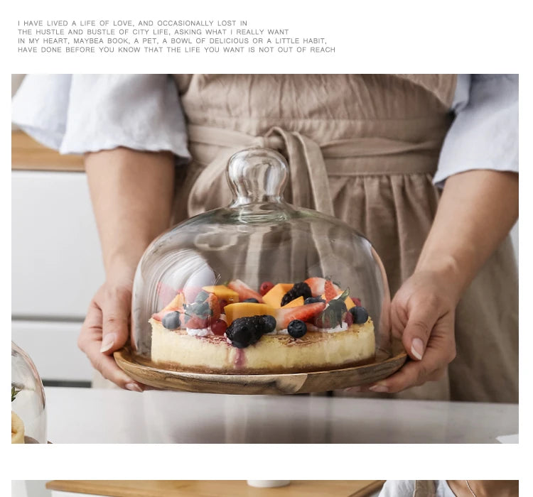 A slice of fruit cake is displayed in a glass dome cover on a wooden tray.