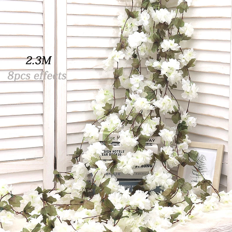 Artificial sakura flower garland with white blossoms and green leaves draped against a white slatted background