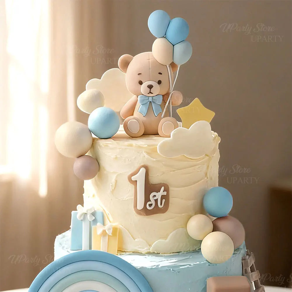 A first birthday cake decorated with a teddy bear topper, blue and white foam balls, and a rainbow design.